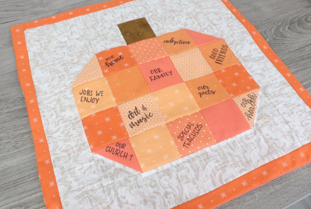 Image is a quilt block composed of an orange patchwork pumpkin with a brown stem on a beige background with an orange border.