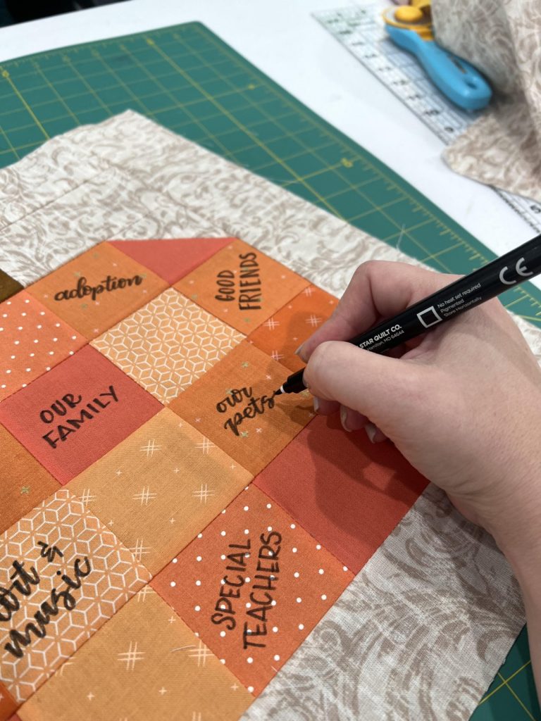 Image contains Amy's hand holding a fabric marker and writing on the pumpkin quilt block.
