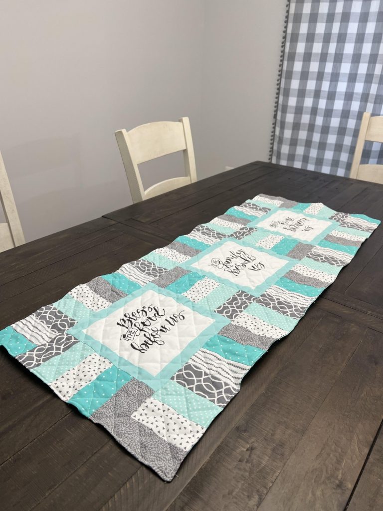 Image contains a grey, teal, and white quilted table runner on a dark wooden table top.