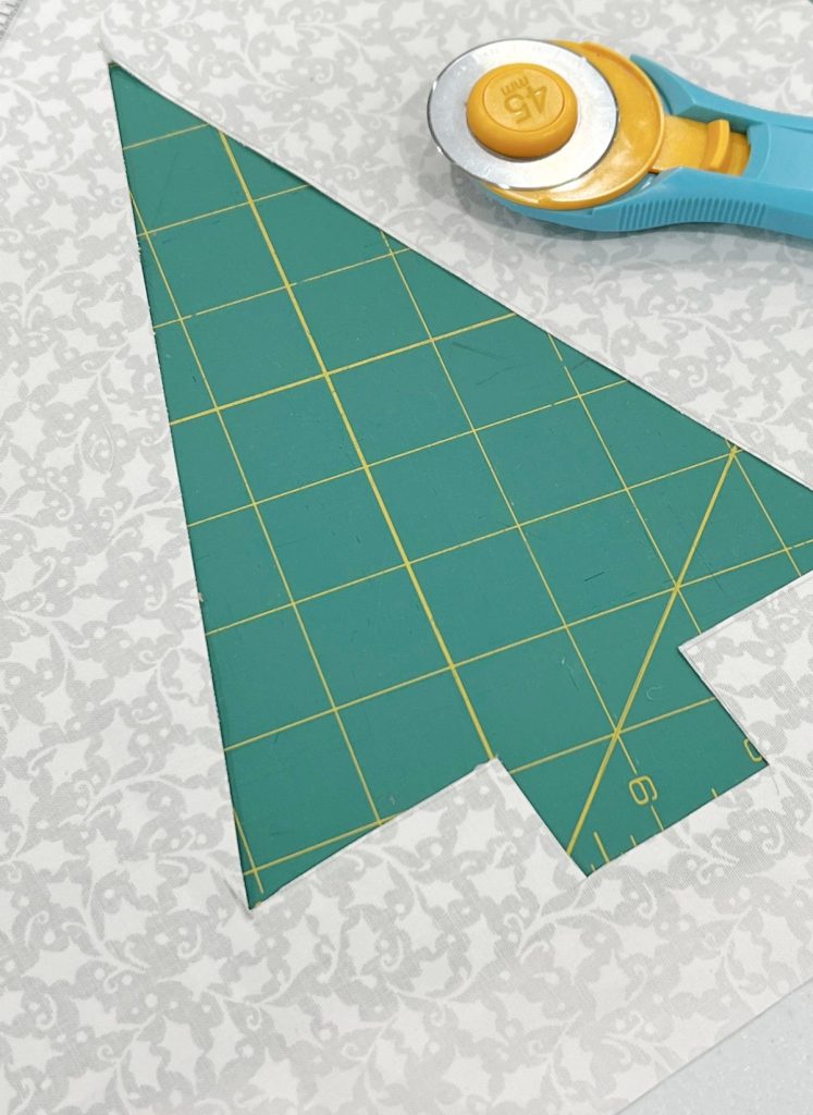 Image contains a piece of white fabric with the shape of a triangle tree cut out of the center. A rotary cutter sits off to the side.
