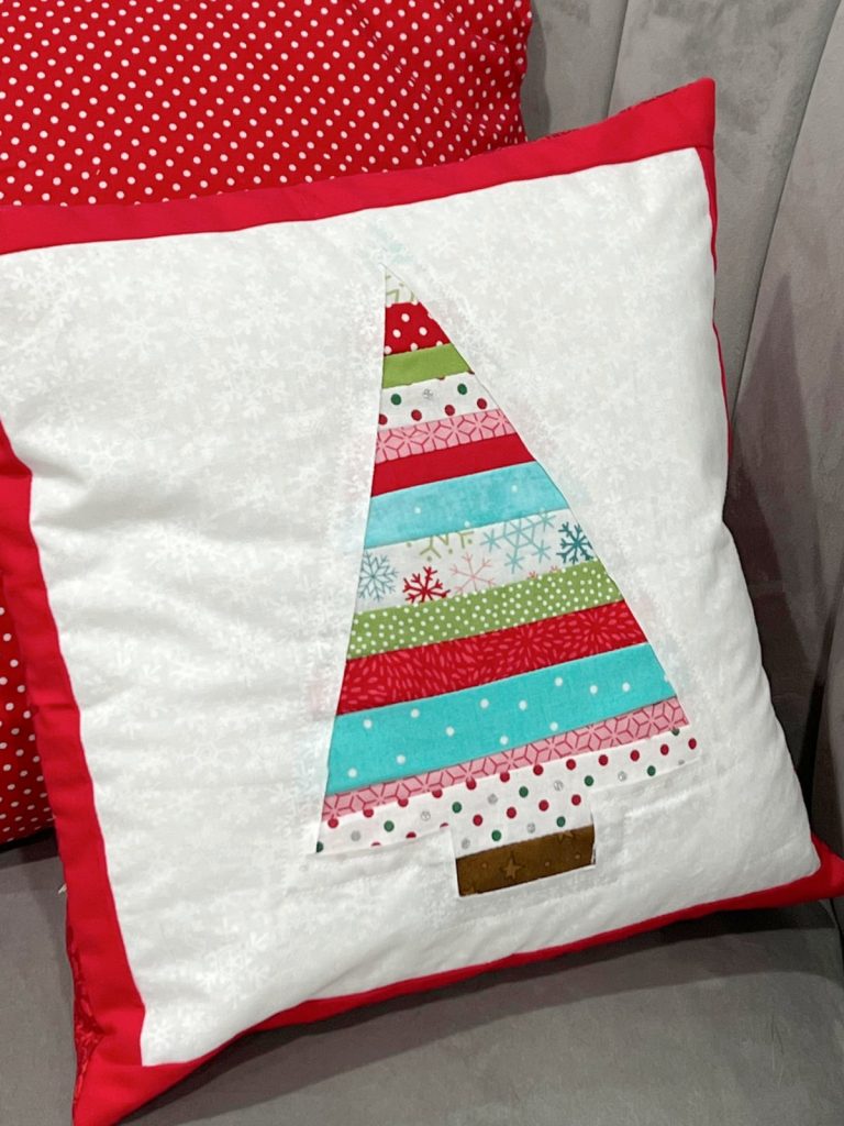 Image contains a white pillow with a multicolored striped tree in the center and a red border, sitting on a grey chair.