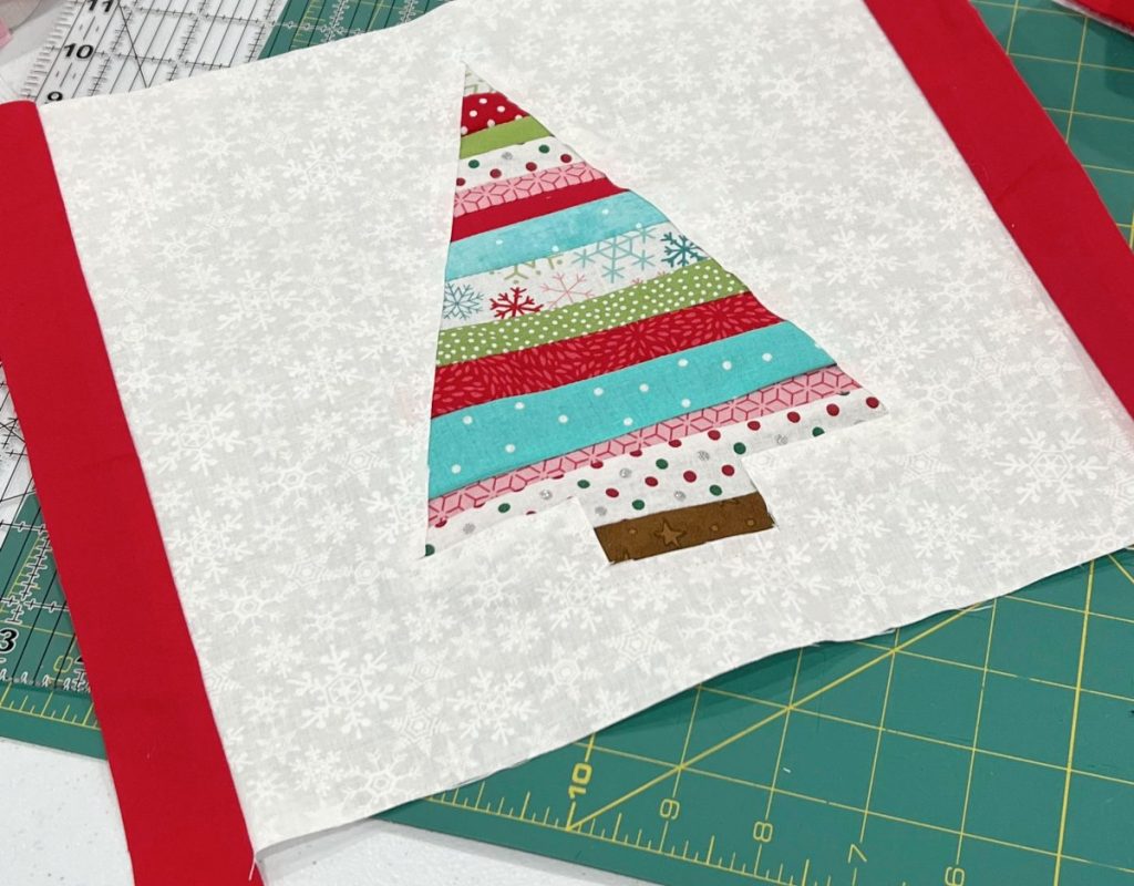 Image contains a white fabric square with a colorful striped tree in the center and red strips on either side.