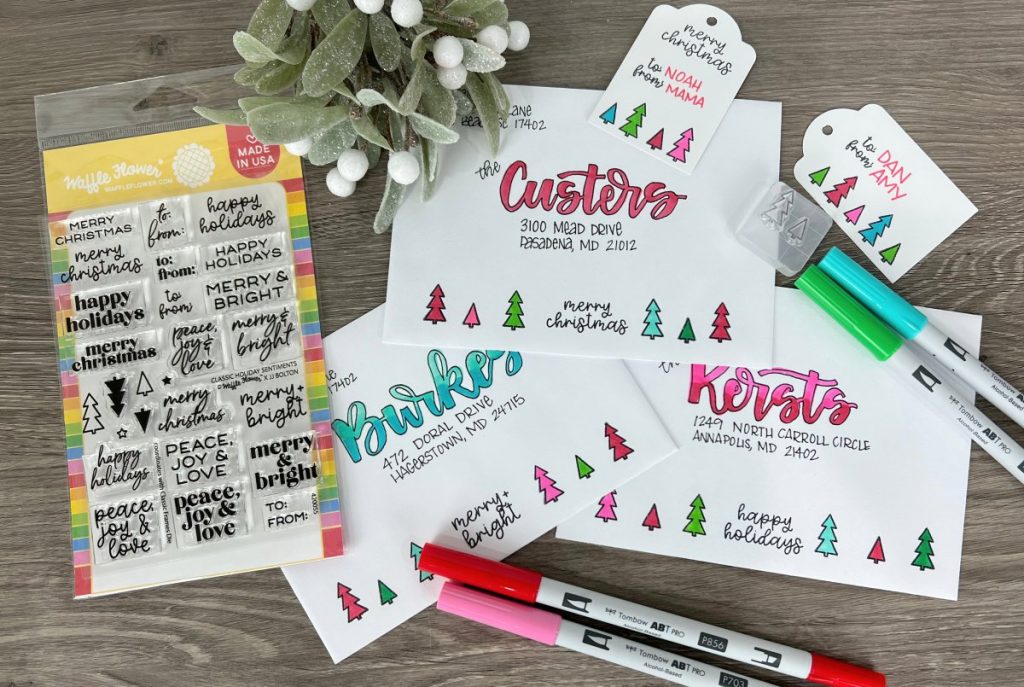 Image contains white envelopes and gift tags stamped with colorful trees. They are surrounded by multicolored markers, a stamp set, and faux mistletoe on a wooden background.
