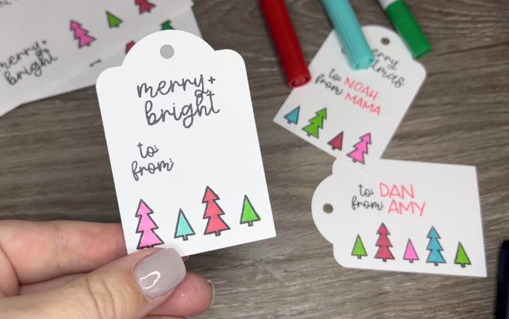 Image contains Amy's hand holding a stamped gift tag that says, "merry and bright" and is decorated with multicolored tr3ees. Other gift tags and markers are in the background on a wooden desktop.