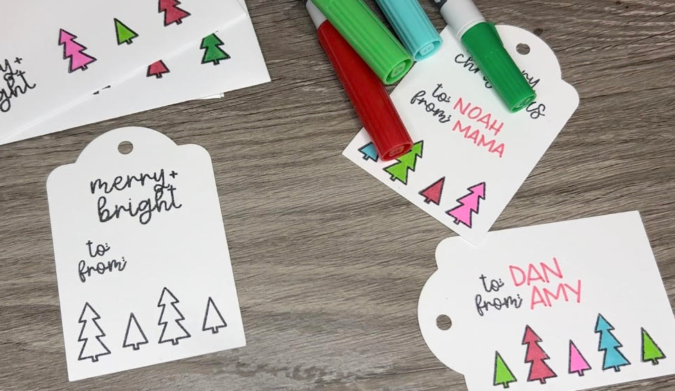 Image contains three stamped white gift tags with trees on them. They sit on a wooden background with four colorful markers.