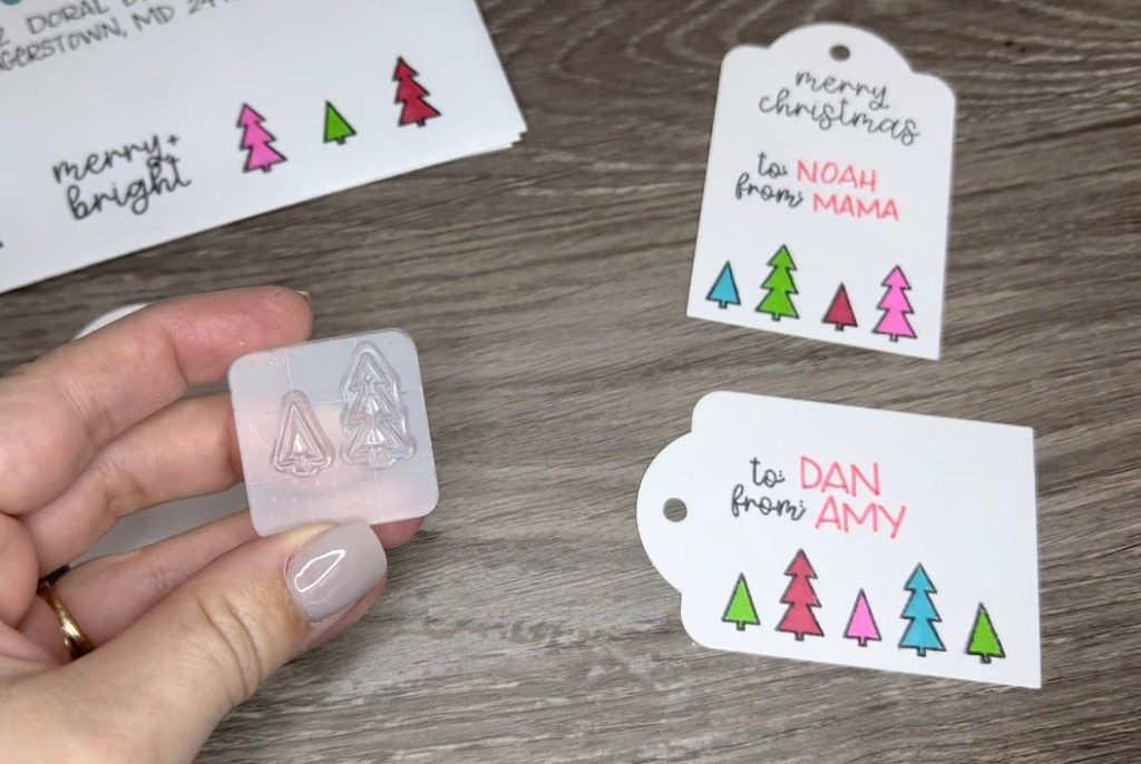Image contains Amy's hand holding an acrylic block with two small tree stamps attached. In the background are two stamped gift tags & a stamped envelope.