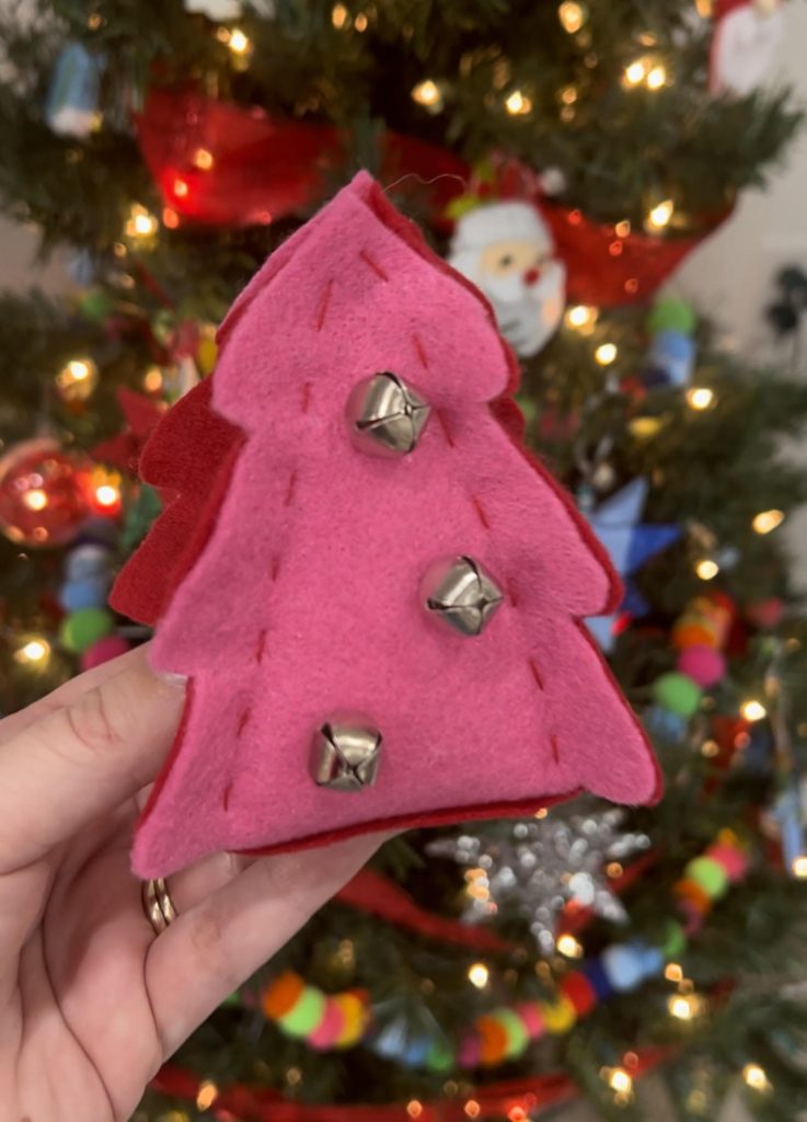 Image contains Amy's hand holding a pink and red felt tree decorated with three silver jingle bells in front of a decorated Christmas tree.