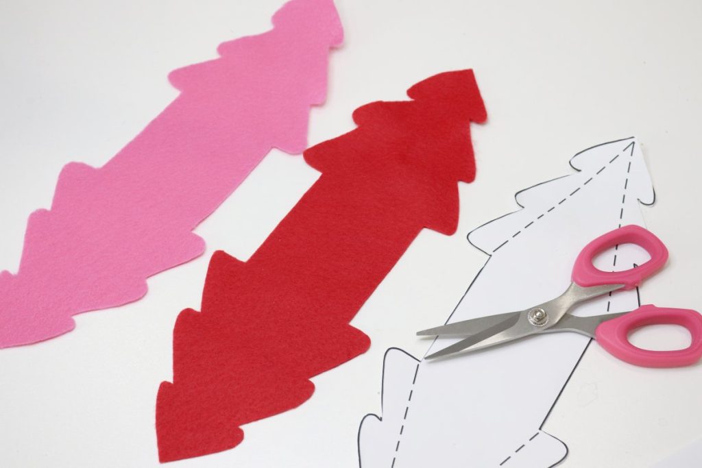 Image contains a pink piece of felt and a red piece of felt cut into the tree template shapes. The paper template sits beside them with a pair of pink-handled scissors on top.