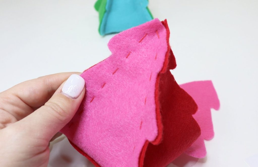 Image contains Amy's hand holding the cut felt pieces for a pink and red tree, partially stitched together with red thread. A teal and green tree is in the background.