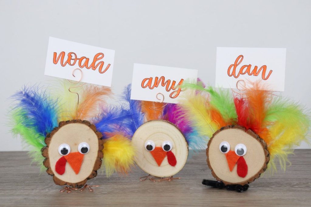 Image contains three wood slice turkeys on a wooden table. Each has a place card attached with a name written on it in orange marker.