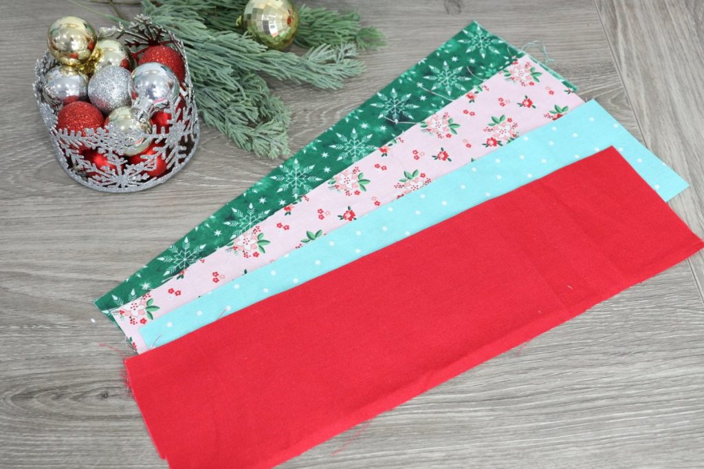 Image contains four strips of fabric; green, pink, teal, and red, on a wooden background with a pine branch and a silver container of ornaments in the background.
