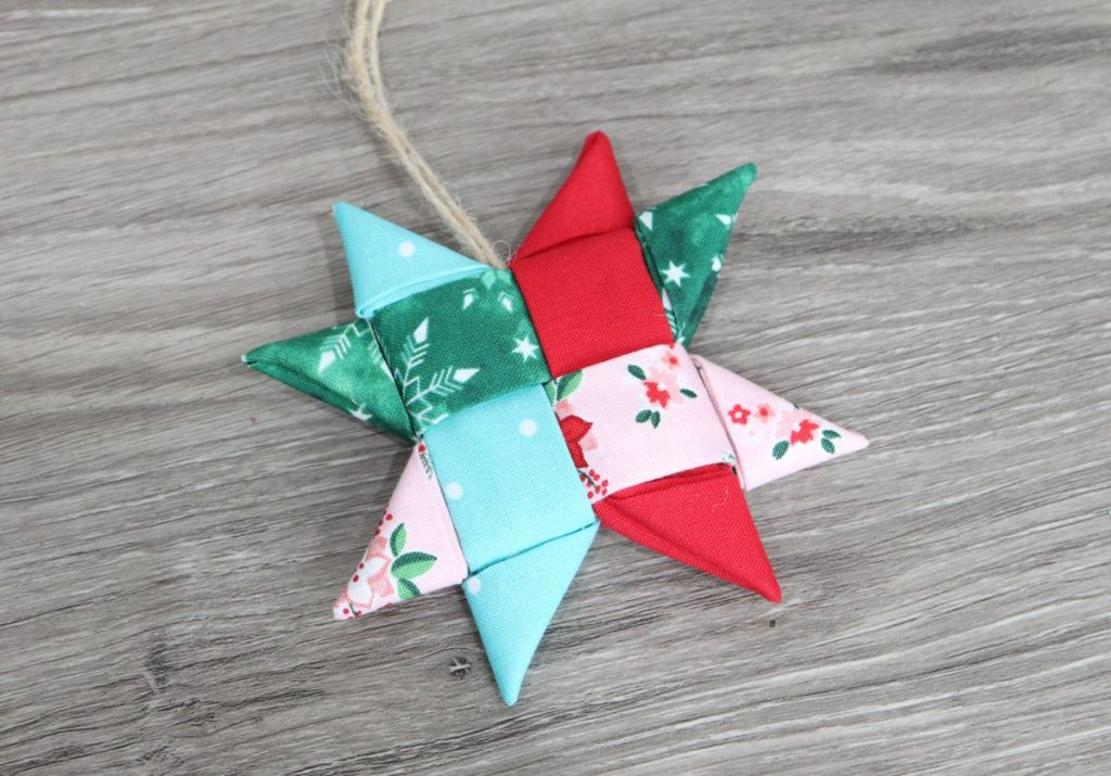 Image contains a folded star ornament made of teal, red, green, and pink fabrics on a wooden background.