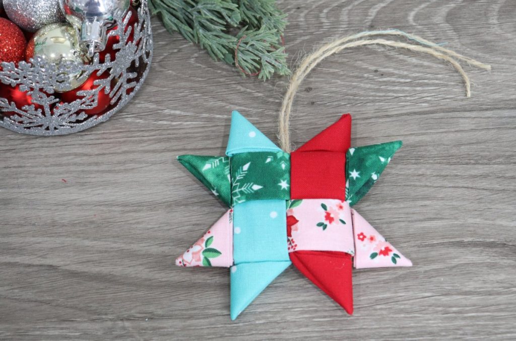 Image contains a folded star ornament made of teal, red, green, and pink fabric, on a wooden background with a pine branch and silver container of ornaments.