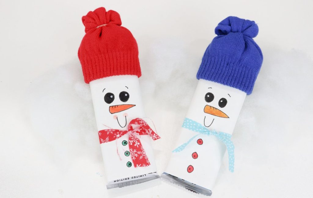 Image contains two candy bars wrapped with white paper and decorated to look like snowmen. One has a red hat and scarf, and the other has a blue hat and scarf.