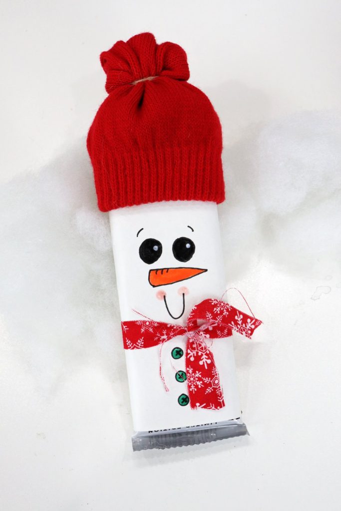 Image contains a candy bar wrapped with white paper and decorated to look like a snowman with a hat made from a red glove and a red fabric scrap scarf.