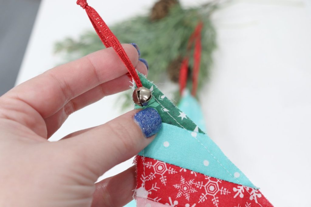 Image contains Amy's hand holding the top of a Scrappy Tree ornament, showing the red ribbon hanger and small jingle bell at the top.