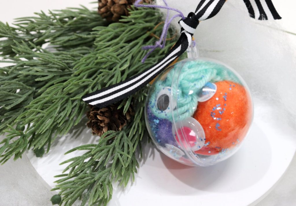 Image contains a clear plastic ornament filled with assorted craft supplies including an orange pom pom, google eyes, blue glitter, and teal string. It sits on a white background with a faux pine branch.