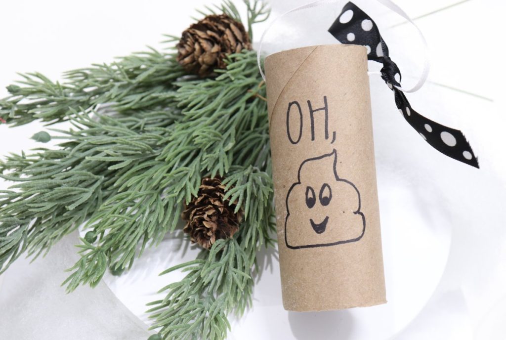 Image contains an empty toilet paper roll with a black and white polka dot bow on top and "OH POOP" in black marker." It sits on a white background next to faux pine branches and pinecones.