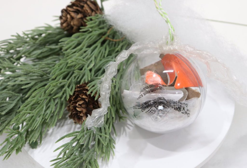 Image contains a clear plastic ornament filled with bits of trash, like pieces of plastic bag and cardboard, on a white background next to faux pine branches.