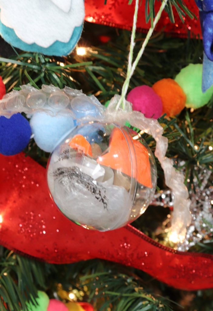 Image contains a clear plastic ornament filled with assorted bits of cardboard and plastic bag, hanging on a decorated tree.