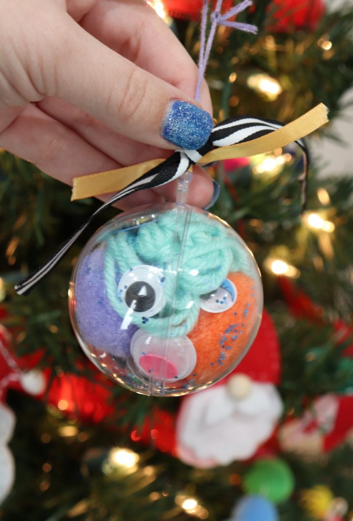 Image contains a clear plastic ornament filled with assorted craft supplies including purple and orange pom poms, google eyes, teal ribbon, and blue glitter, held in front of a Christmas tree.