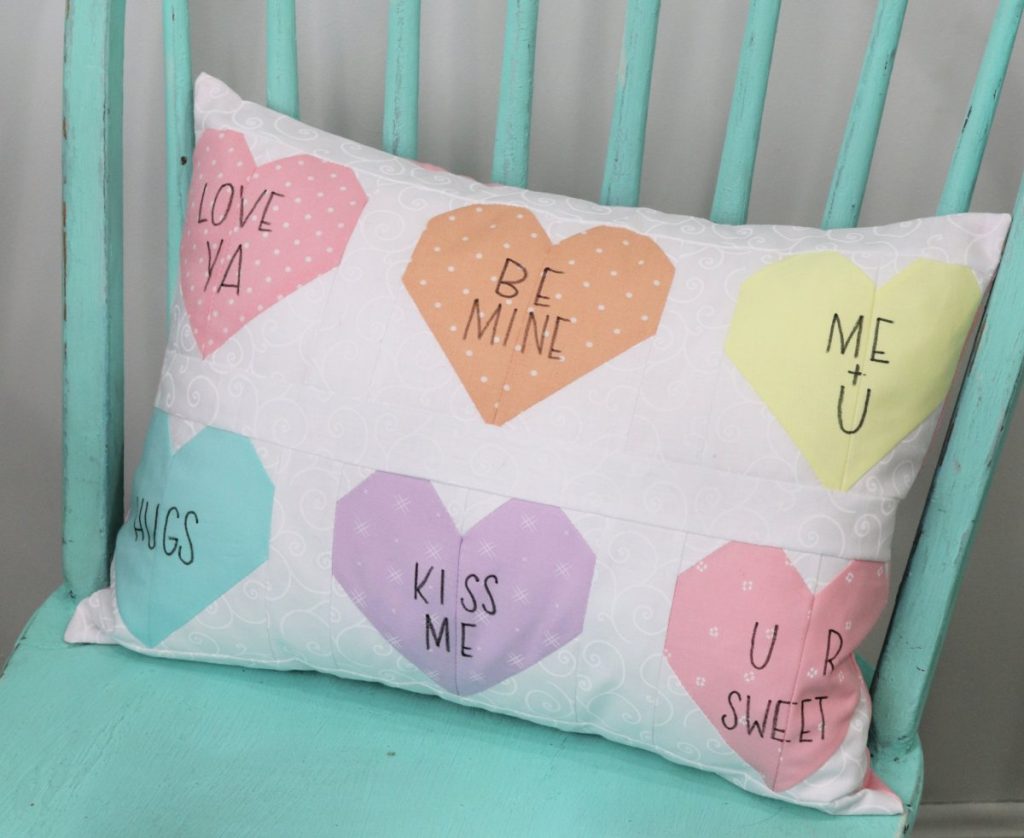 Image contains a white rectangle pillow with six colored fabric hearts on it. Each heart has a message written in black marker, like "love ya," or "be mine."