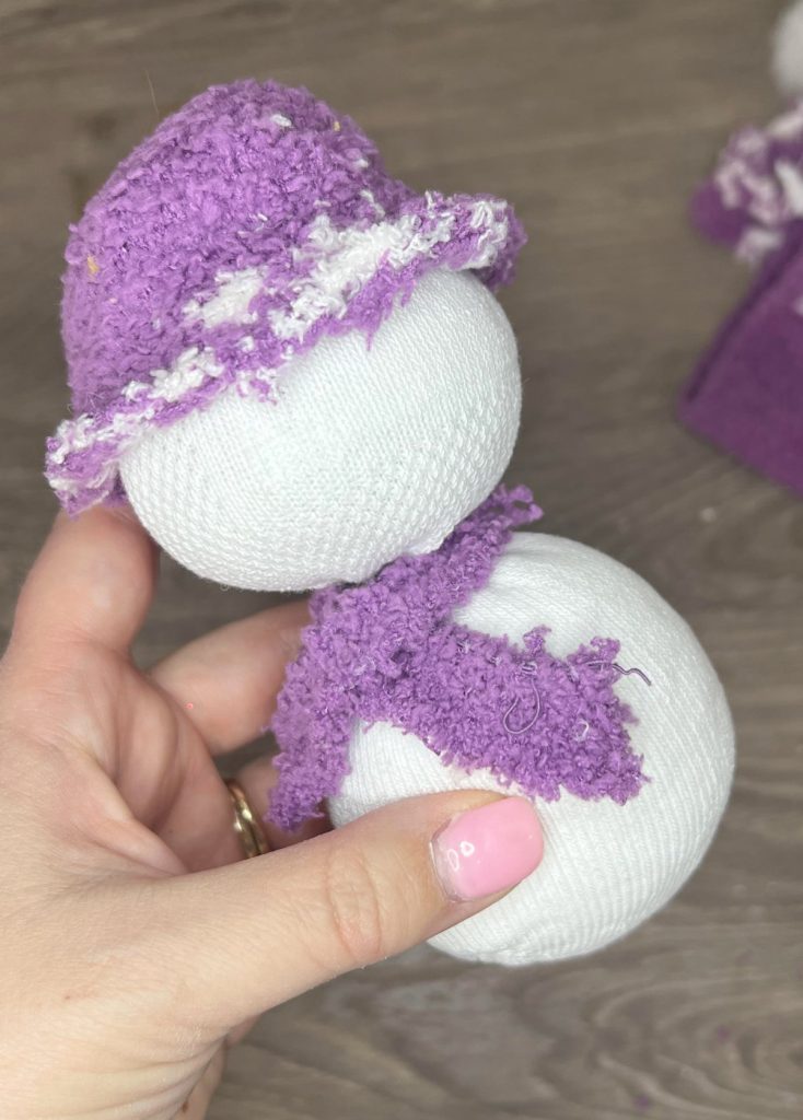 Image contains Amy's hand holding a sock that has been stuffed and formed into two balls, and is wearing a purple fleece hat and scarf.