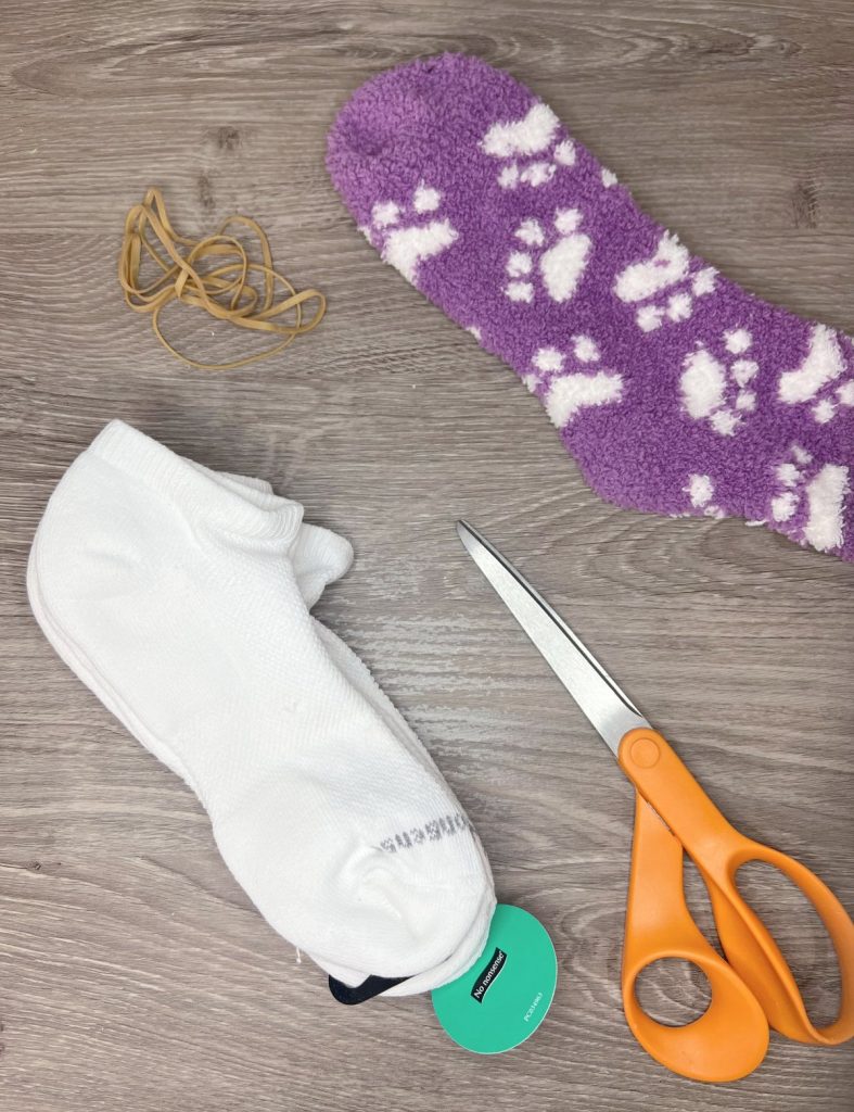 Image contains several white socks, a fuzzy purple sock with white paw prints, several rubber bands, and a pair of orange handled scissors sitting on a wooden table top.