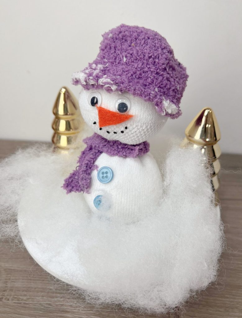 Image contains a snowman made from a white sock with a purple hat and scarf, google eyes, and blue buttons. It is surrounded by white Poly-Fil and gold metallic trees on a wooden table top.