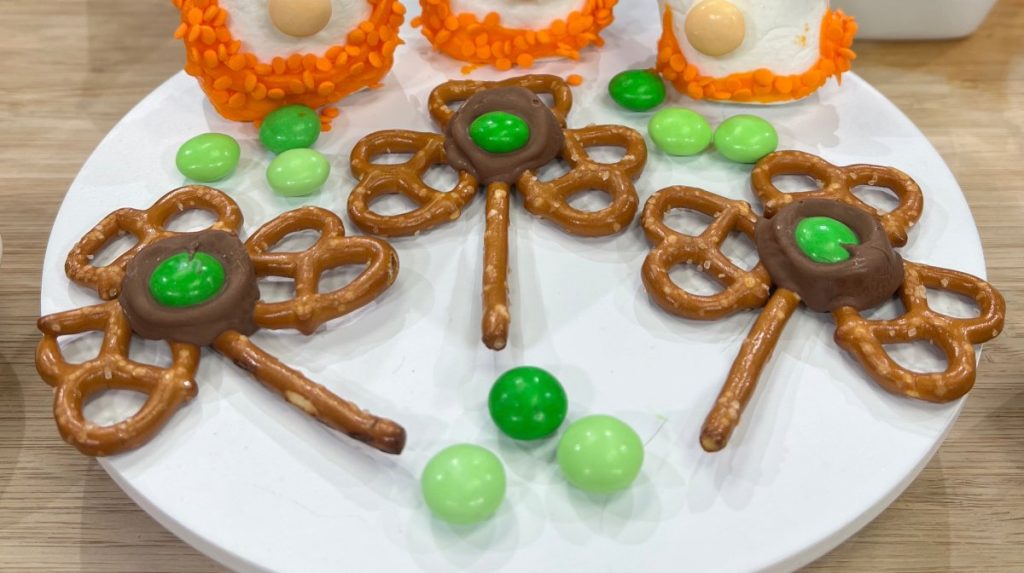 Image contains three pretzel shamrock snacks made by placing three small pretzels and a pretzel stick in a shamrock shape and attaching them together with melted chocolate and a green candy.