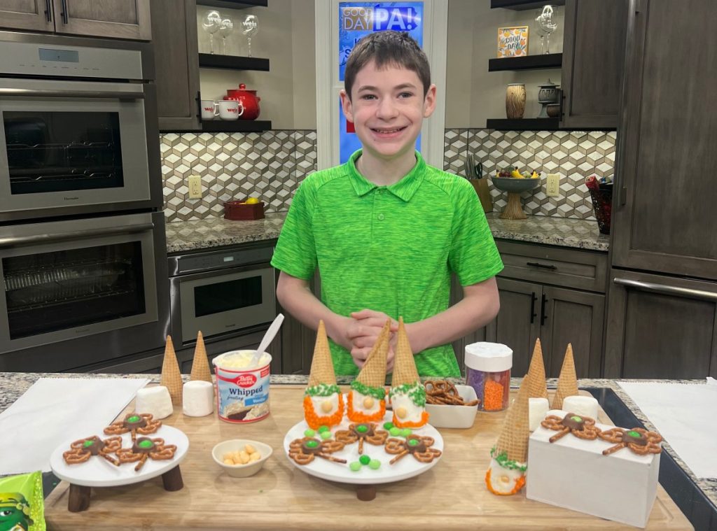 Image contains a smiling boy in a lime green polo shirt standing in a kitchen behind a tray of St. Patrick’s Day treats.
