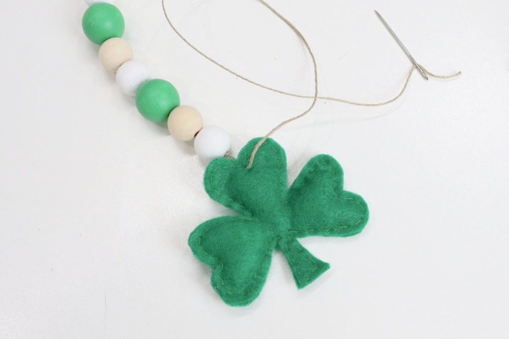Image contains a plush green felt shamrock being attached to the end of a wooden bead garland with a tapestry needle.