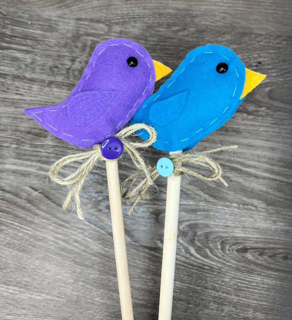 Image contains two felt birds; one purple and one blue, on top of wooden dowels decorated with twine bows and coordinating buttons.