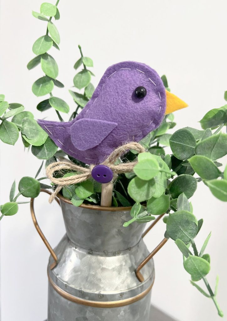 Image contains a purple felt bird on a wooden dowel with a decorative twine bow and purple button. It sits in a tin milk jug filled with faux eucalyptus.