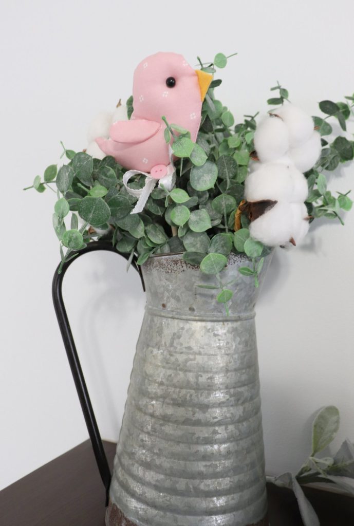 Image contains a plush pink bird on a wooden dowel in an arrangement of faux eucalyptus and cotton.