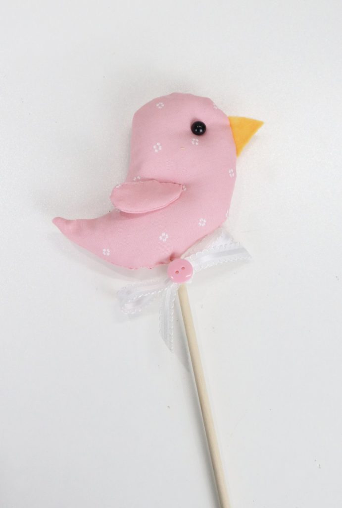 Image contains a small plush bird made of pink fabric sitting on top of a thin wooden dowel.