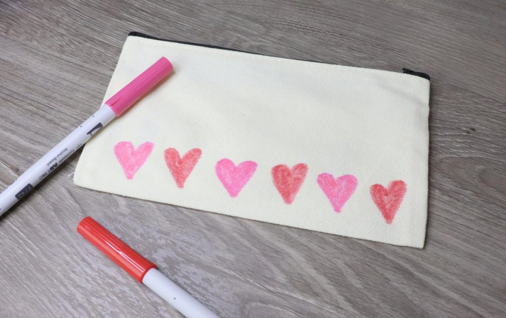 Image contains a cream colored pouch with red and pink hearts across the bottom, and red and pink markers nearby.