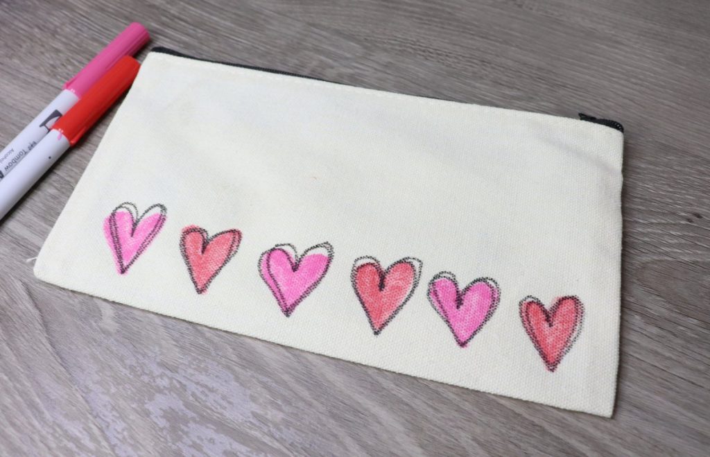 Image contains a cream colored pouch with red and pink hearts across the bottom, and two markers off to the side.