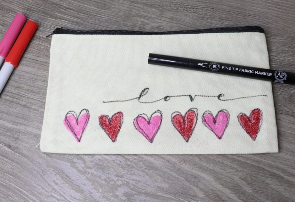 Image contains a cream colored canvas pouch with six red and pink hearts across the bottom and the word "love" written in black script. Markers and a fabric pen lay around it.