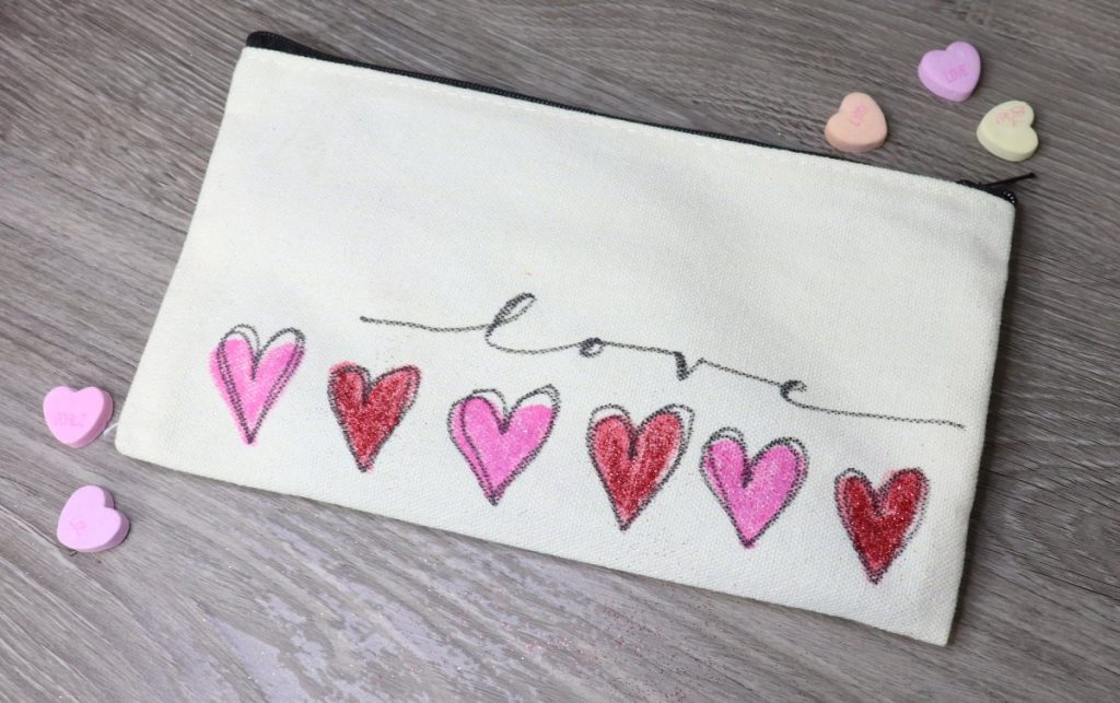 Image contains a cream colored canvas pouch with six red and pink hearts across the bottom and the word "love" written in black script.