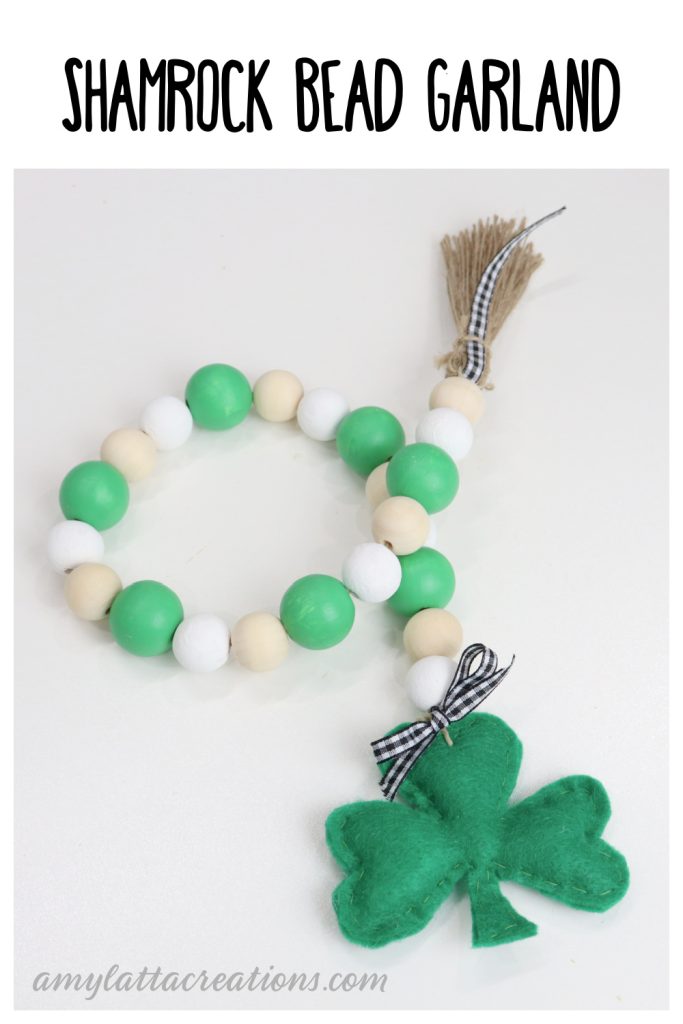 Image contains a decorative garland made of green, white, and natural wooden beads. On one end is a jute tassel, and on the other is a green felt shamrock.