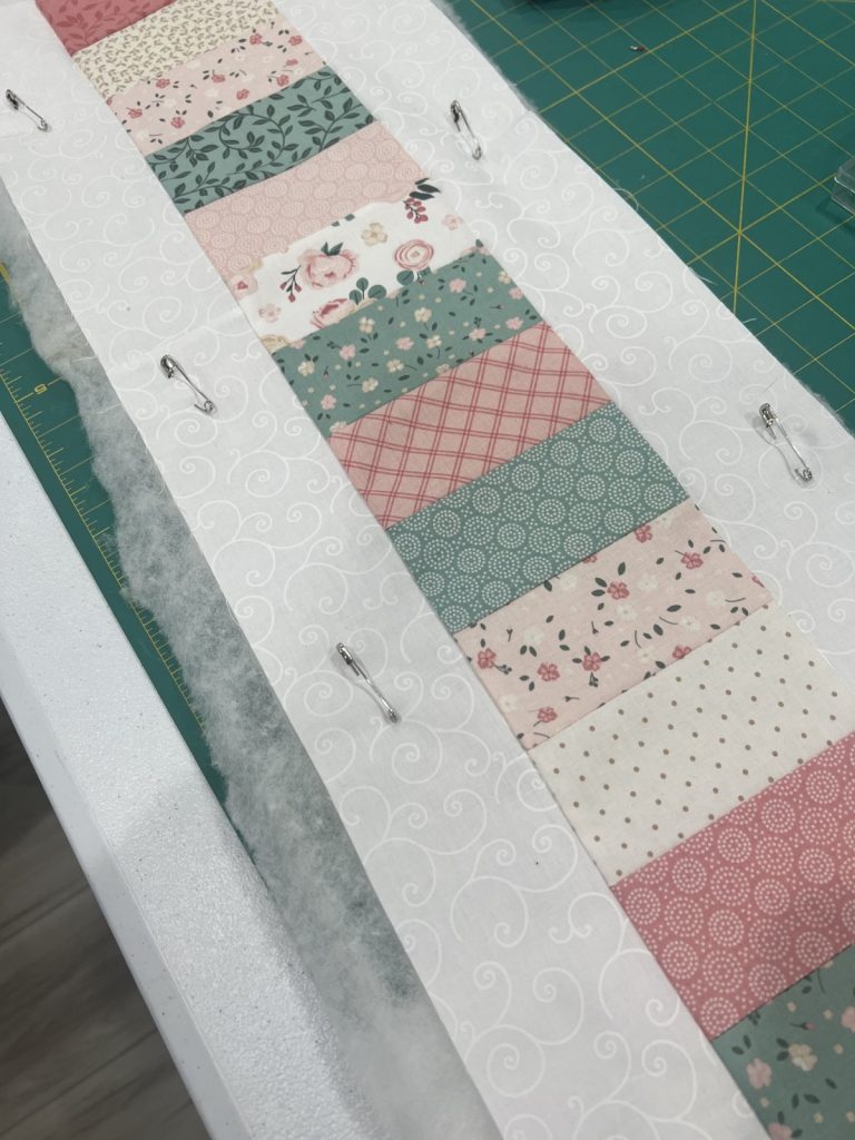 Image contains the top of a table runner pinned to batting and backing fabric.