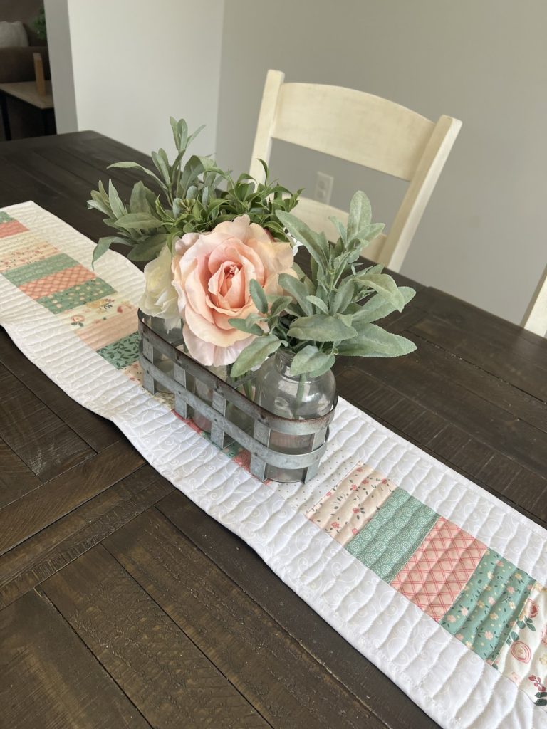 Image contains a wood top table with a quilted table runner and a floral centerpiece.