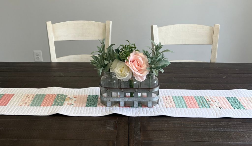 Image contains a quilted table runner on top of a wooden dining room table with a floral centerpiece.