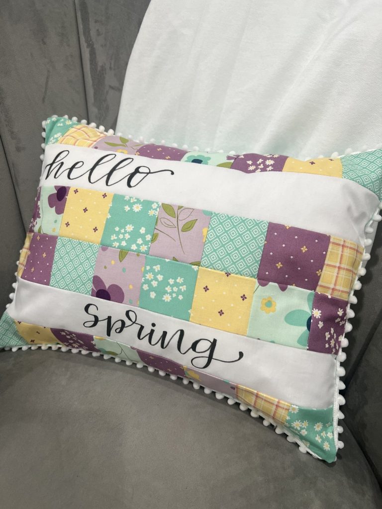 Image contains a patchwork pillow made from pink, teal, and yellow fabric with the words “hello spring” written on it in script.