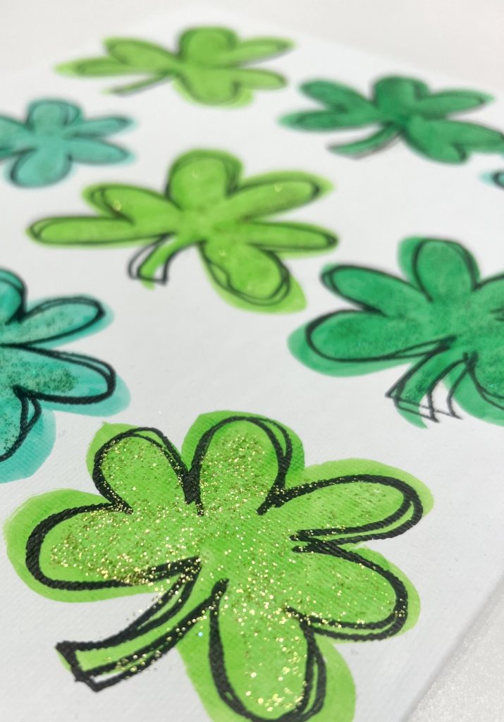 Image is a closeup of a green shamrock drawn on a white canvas with green glitter on top.
