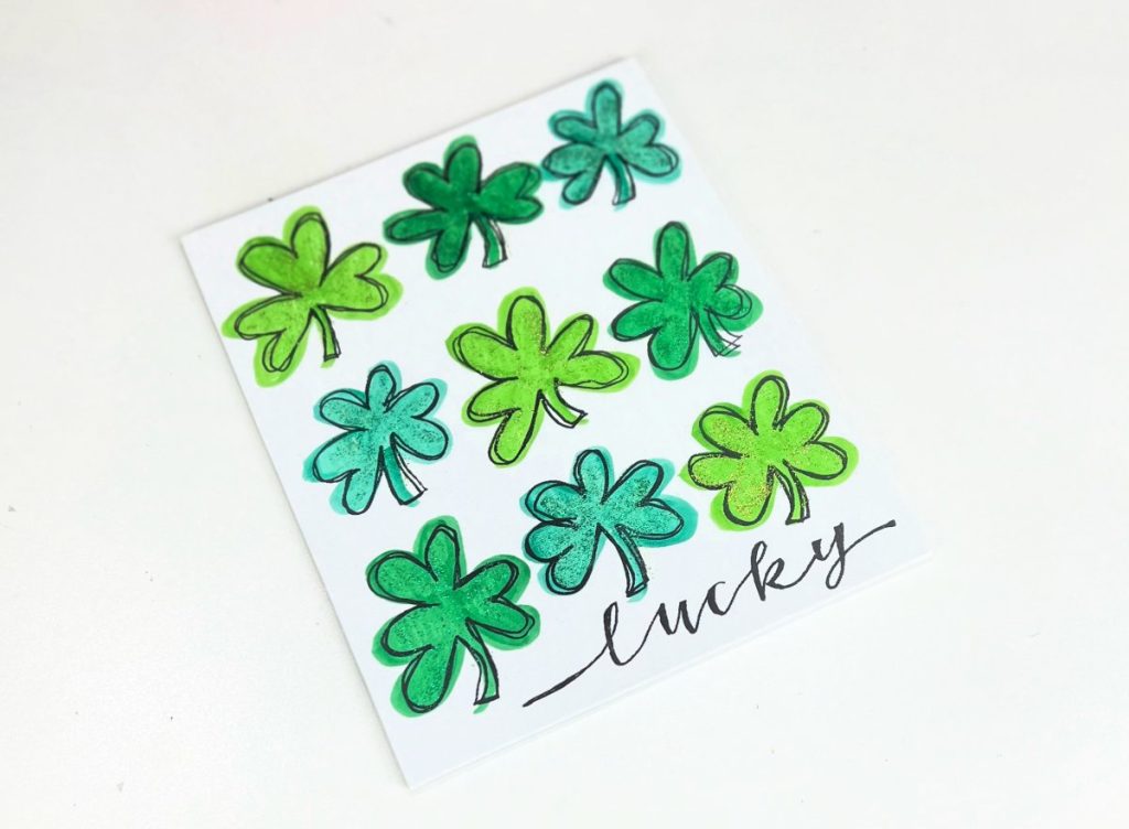 Image contains a white canvas with nine green shamrocks drawn on it and the word, “lucky” written in black script.
