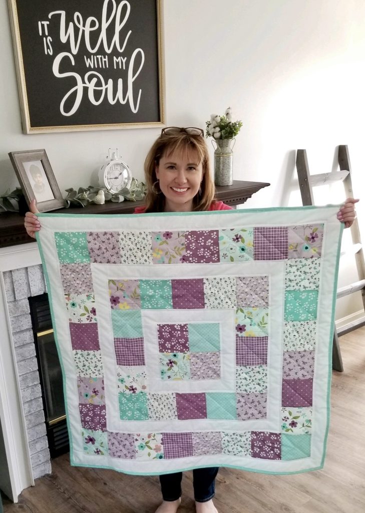 Image contains a smiling woman holding a square quilt made from teal, purple, and white fabric squares.