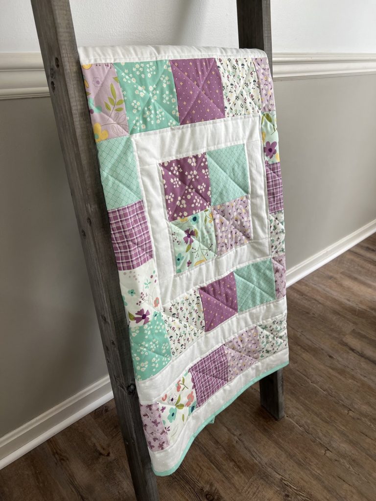 Image contains a quilt made from purple, teal, and white floral squares.