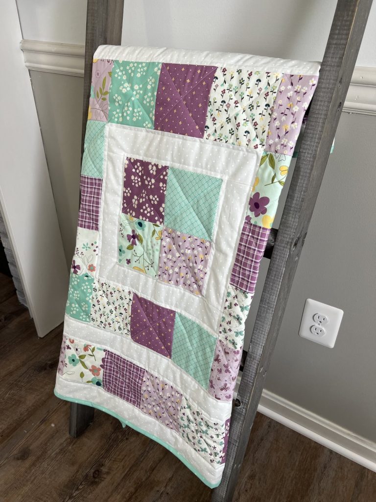 Image contains a quilt made from white, teal, and purple floral fabric squares, displayed on a grey wooden ladder.